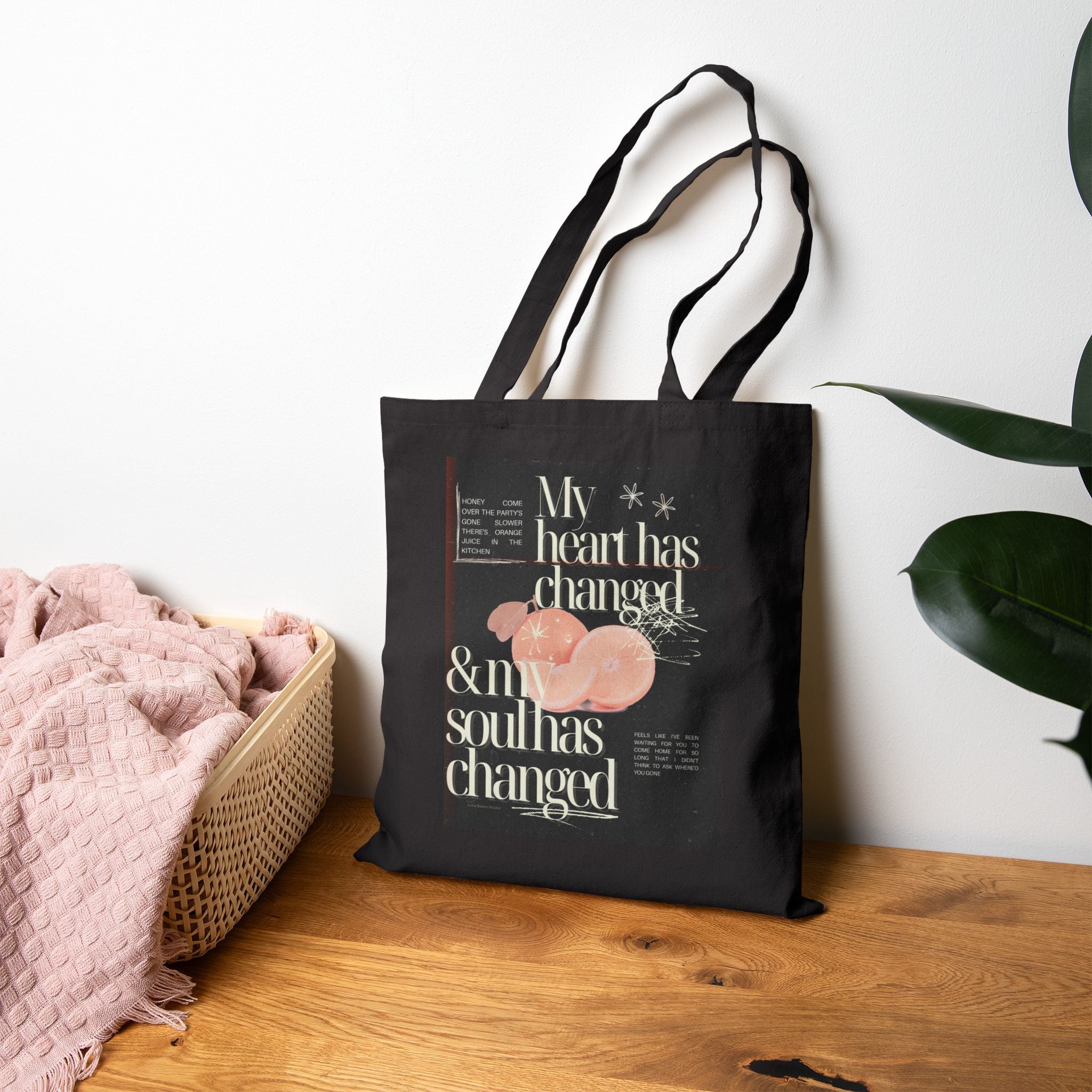 Black tote bag with text and design on a wooden surface with a white wall and plant in the background